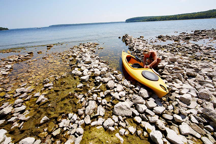 A kayaker in water near a beach of rocks.