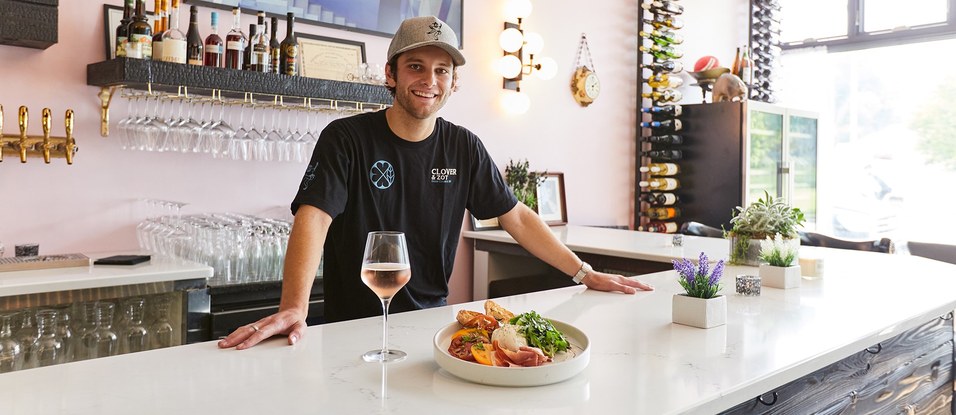 Man standing behind a counter with a plate of food and a glass of white wine in front.