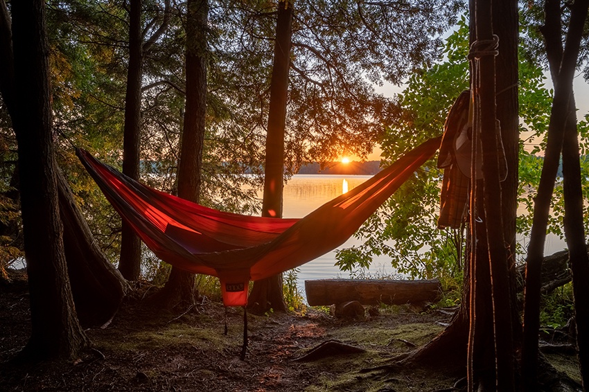 A hammock hangs between two trees at a lakeside campsite at sunset.