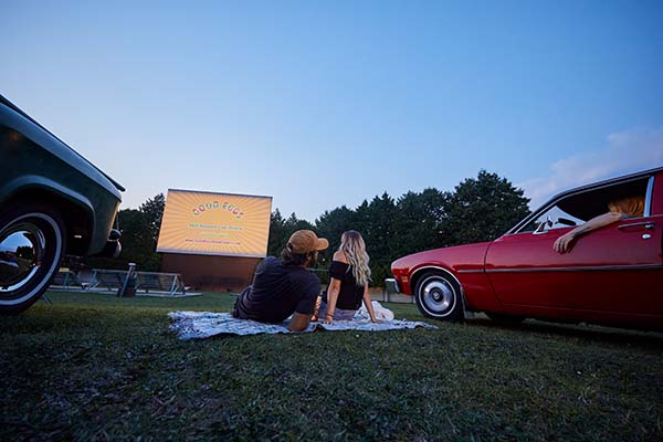 A couple sits on a blanket on the grass aside their car the drive-in movie theater.